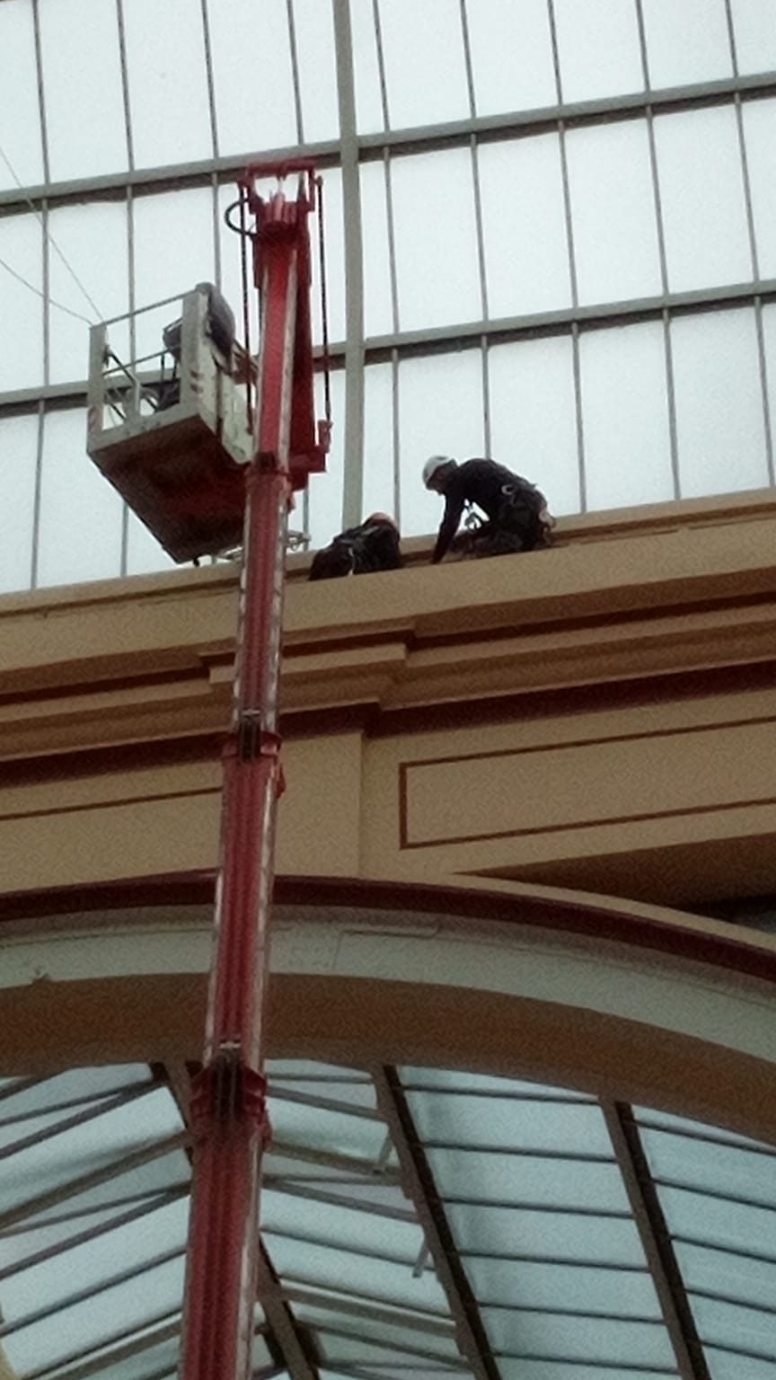 Two technicians on a high interior ledge with a red spider boom in the glazed atrium at Alexandra Palace