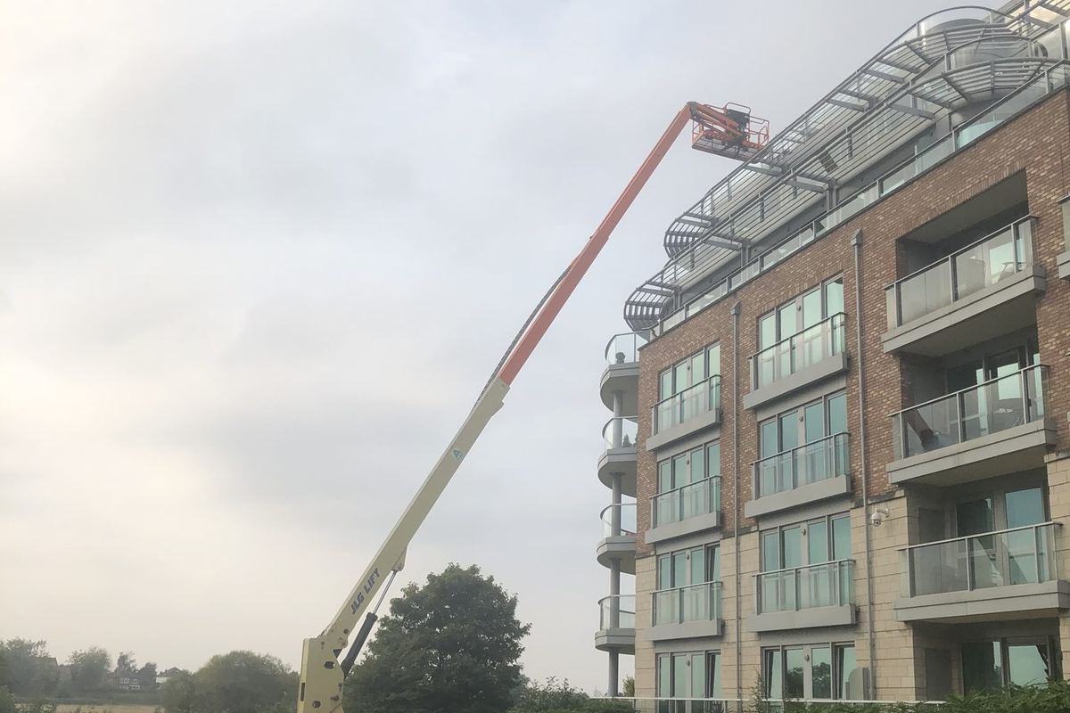 Orange boom lift raised to roof level beside a multi-storey block, giving glaziers access to the high-level glazing and roof canopy