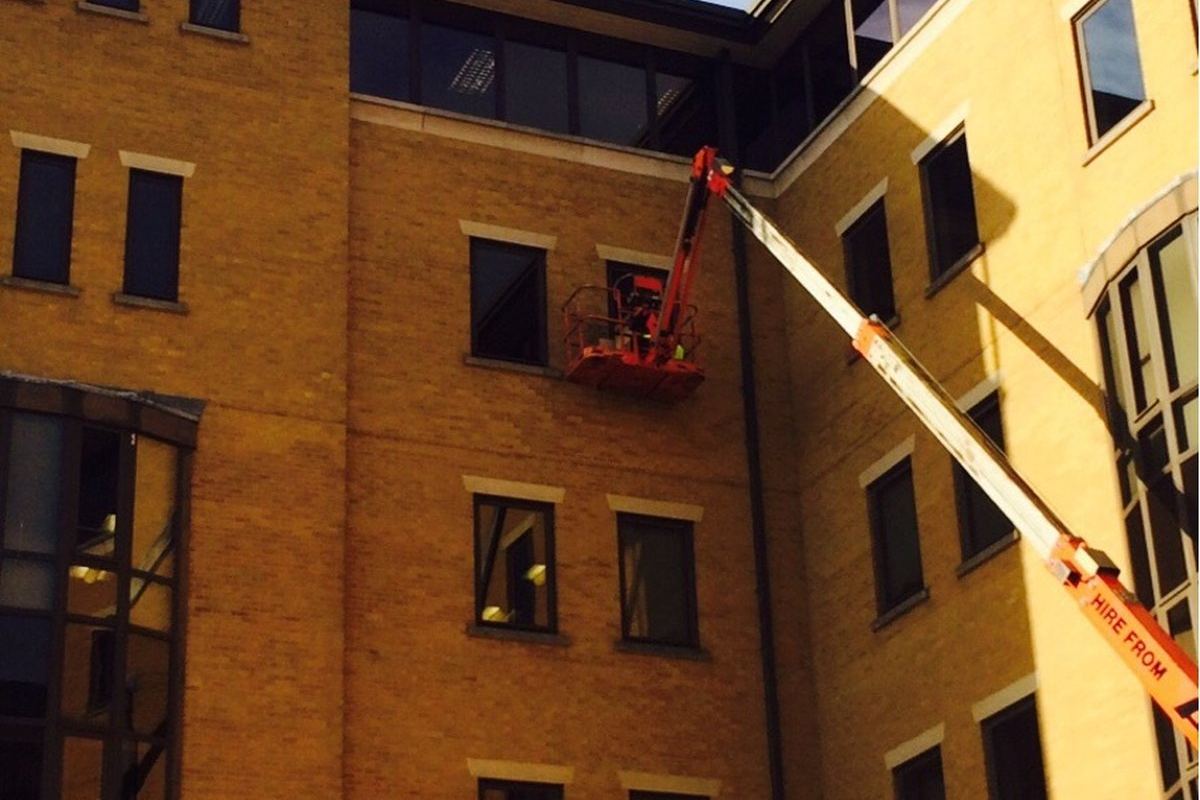 Boom lift raised against a yellow-brick office building with a glazier in the basket at an upper-floor window, replacing glass without scaffolding