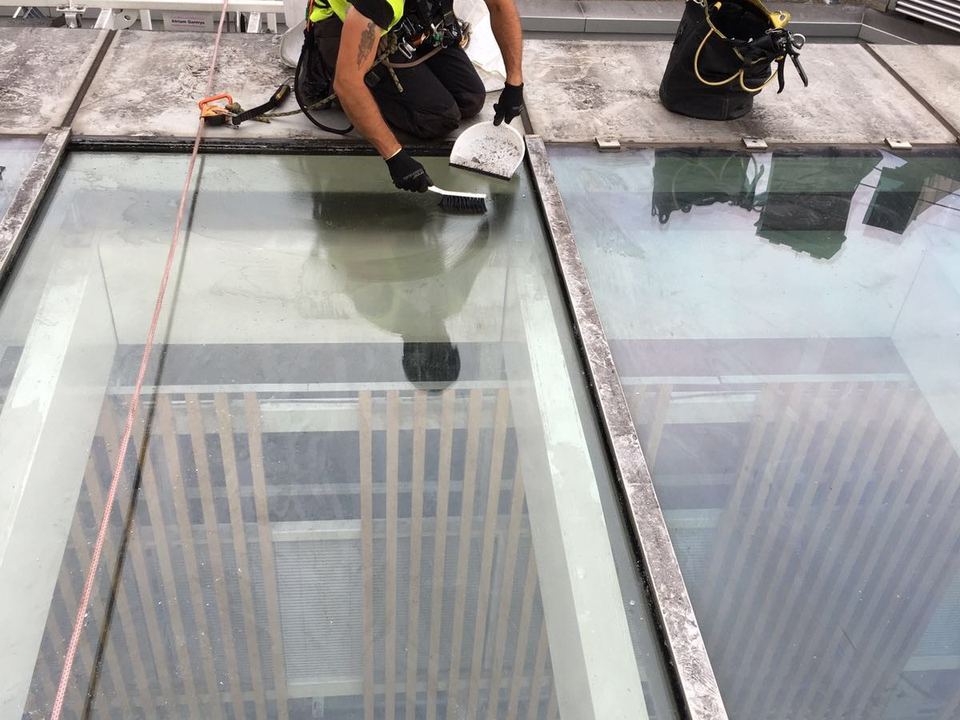 Rope-access technician in a harness, tied off to a rope line, working on the seals of a sloped glass roof