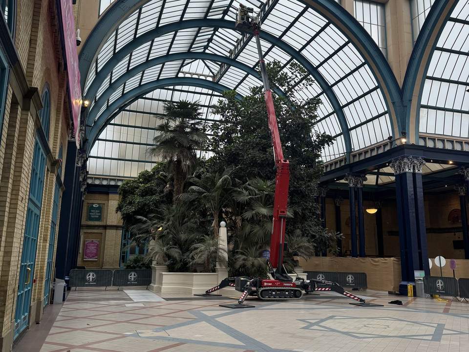 Tracked spider boom lift set up on a protected floor inside a glazed atrium with site barriers, reaching the glass roof without scaffolding